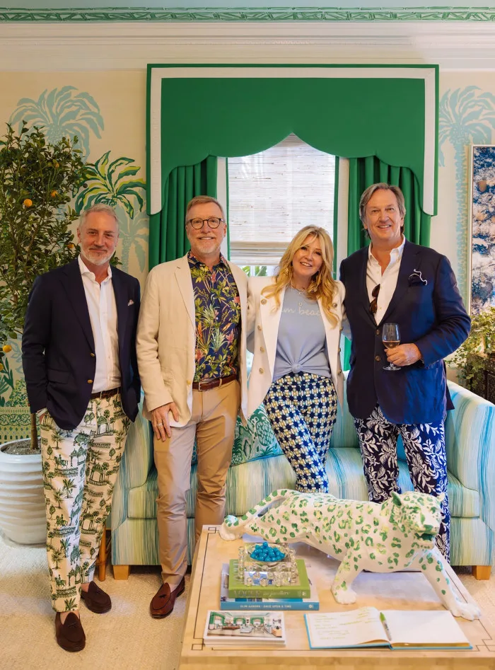 Four people stand in a room decorated with green and blue patterns, including a woman in patterned pants and a man holding a wine glass.